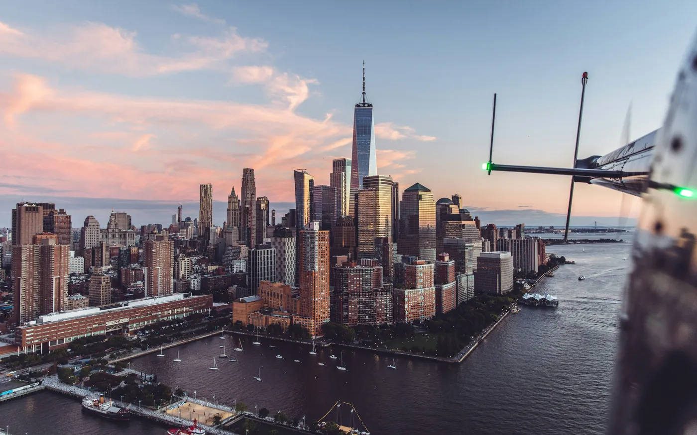 Manhattan skyline with One World Trade Center seen from a helicopter at sunset, with the helicopter skid visible at the edge of the frame